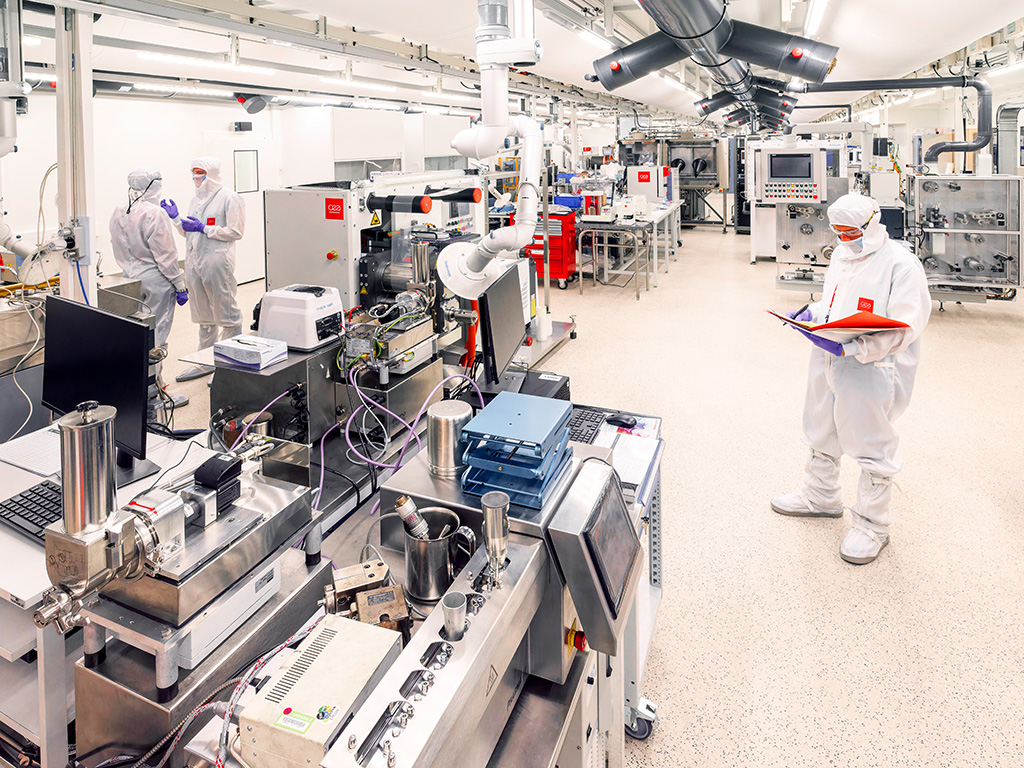 Photo of a clean room with machines and pipes, people in white overalls work in it