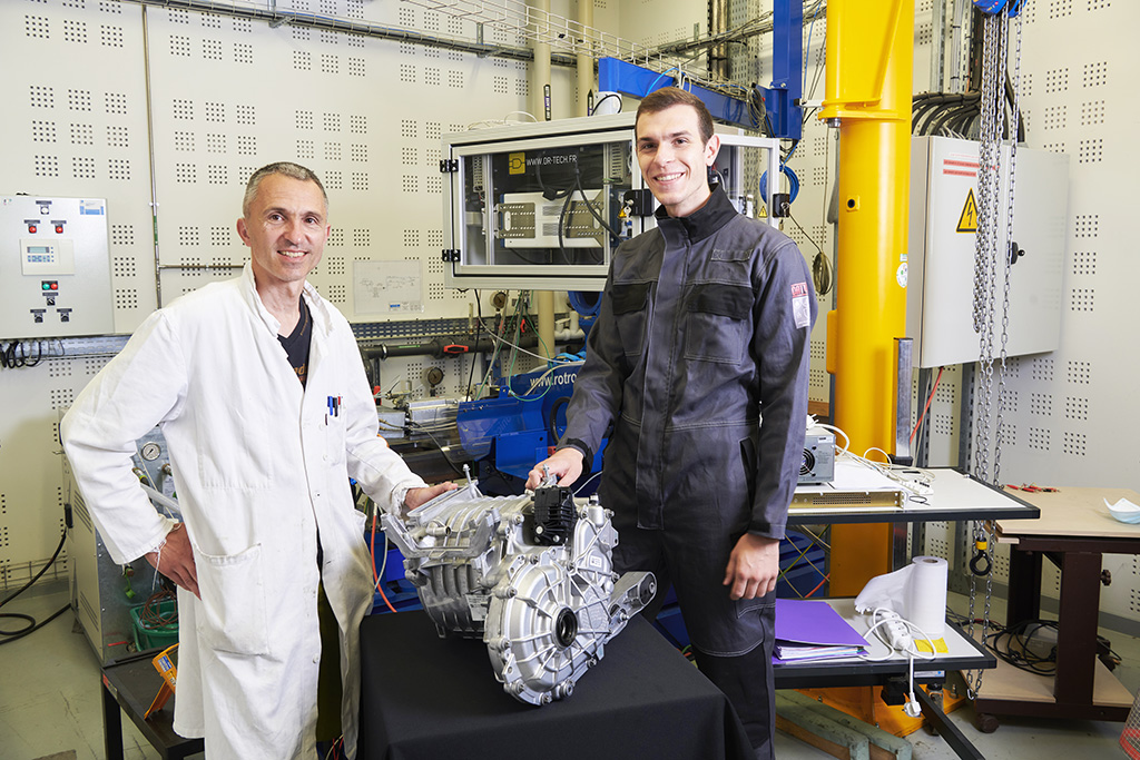 two men, one in laboratory clothing and the other in a pilot's suit, pose with an automobile engine