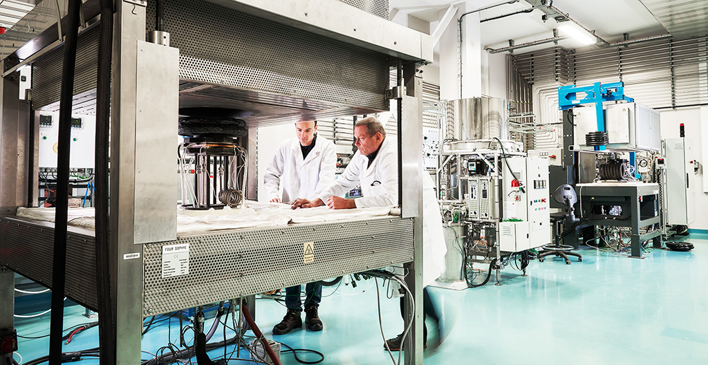 two men in laboratory outfits work in a room with large machines