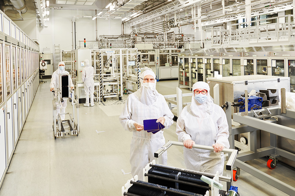 Photo of the interior of a clean room. Three people dressed in white jumpsuits and wearing masks pose with rolling carts. One of the people is holding photovoltaic panels in his hands.