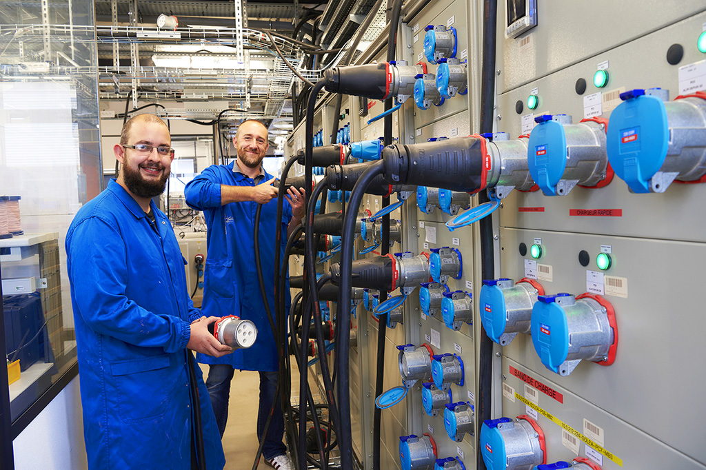 two men dressed in blue overalls pose in front of a wall with giant sockets and cables