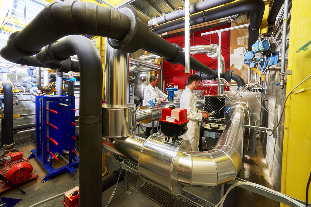 two men in laboratory outfits work among huge pipes